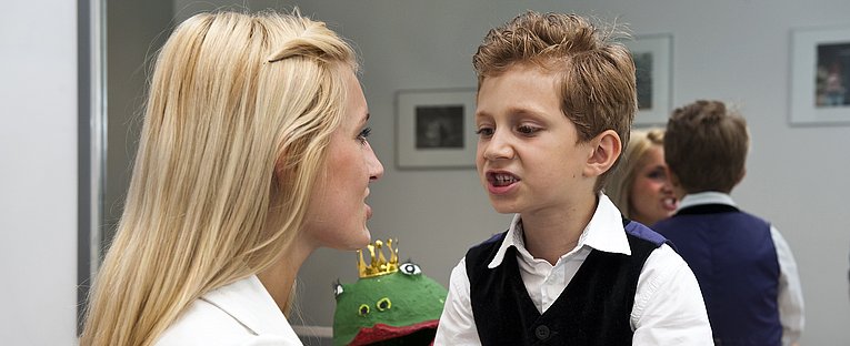Eine blonde Logopädin im Halbprofil von schräg hinten die einem Kind bei einer logopädischen Behandlung gegenübersitzt, das bei zusammengepressten Zähnen den Mund geöffnet hat.